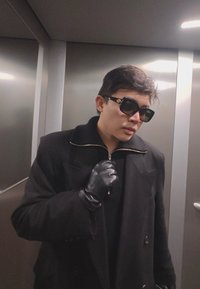 Fiatal férfi fekete kabátban és bőrkesztyűben, aki a gallérját igazítja, nagy fekete napszemüveget visel, egy fémes lift belsejében áll.