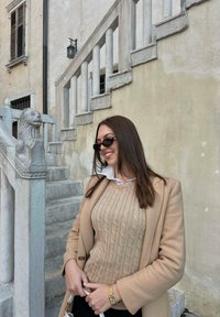Beige svetr s pleteným vzorem v kombinaci s hnědým kabátem, černými kalhotami a slunečními brýlemi, stojící poblí�ž kamenné sochy lva a schodiště.