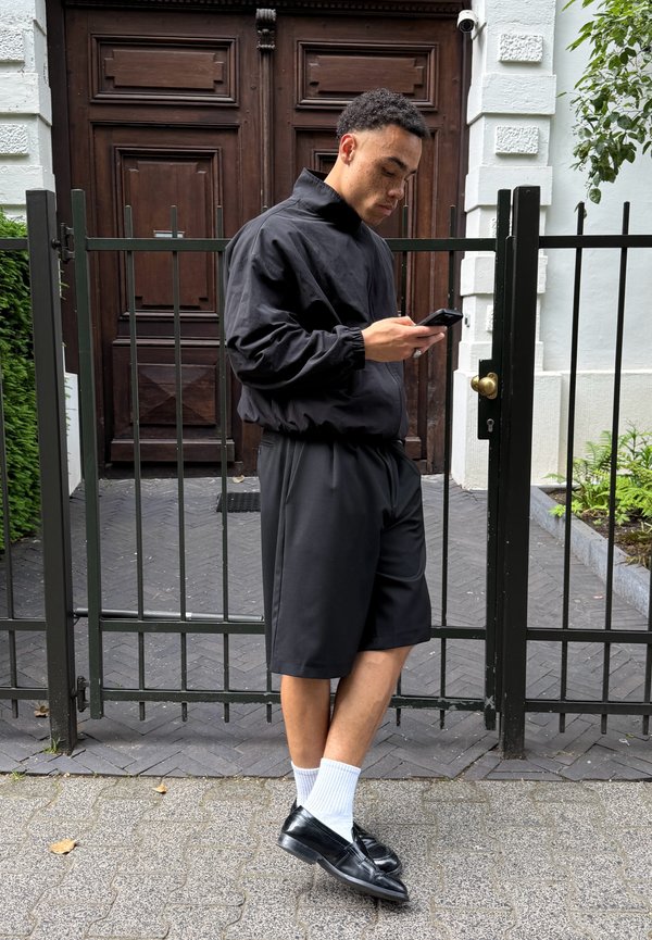Ung mand i sort jakke og shorts står ved en metalport og kigger ned på en smartphone i hænderne.