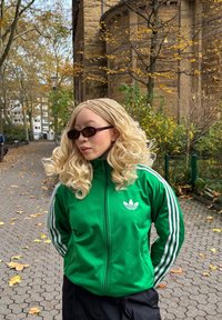 Casaco verde com fecho de correr, riscas brancas e logótipo, usado com calças pretas. Cabelo loiro comprido e ondulado com tranças e óculos de sol escuros. Folhas de outono ao fundo.