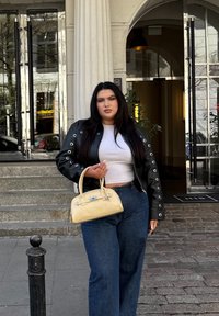 Čierna kožená bunda s očkovými detailami, biele crop top, široké modré džínsy a malá žltá kabelka s modrými akcentmi.