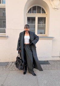 Άτομο που φορά μακρύ σκούρο γκρι παλτό, λευκό τοπ σωλήνα, παντελόνια με ρίγες και πλατύ πόδι, μαύρες μπότες και γάντια, κουβαλώντας μεγάλη μαύρη τσάντα, στέκεται στο πεζοδρόμιο.