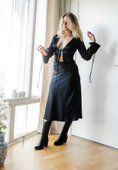 Top negro estilo envolvente con mangas largas y acentos de lazo, combinado con una falda negra a juego. Lleva botas de tacón negras con textura.