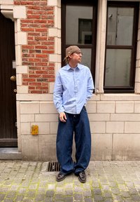 Mies, joka pukeutunut vaaleansiniseen paitaan, ylipitkiin tummansinisiin housuihin, ruskeisiin kenkiin ja kuvioituun lippalakkiin, seisoo mukulakivikadulla lähellä tiili- ja kivirakennusta.