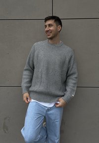 Sivý hrubý pletený sveter s rebrovanou textúrou a okrúhlym výstrihom, v kombinácii so svetlomodrými džínsami. V pozadí je hladká sivá stena.