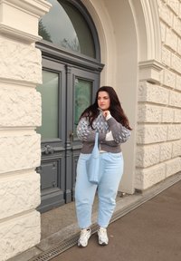 Grå og blå mønstret genser, lyseblå jeans og hvite joggesko. Holder en lyseblå håndveske, står nær en strukturert steinvegg.