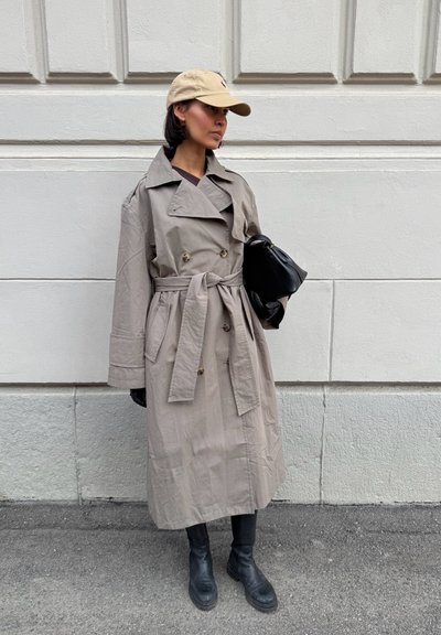 Mujer con gabardina beige, gorra beige, botas negras y un bolso negro, de pie contra una pared gris claro.