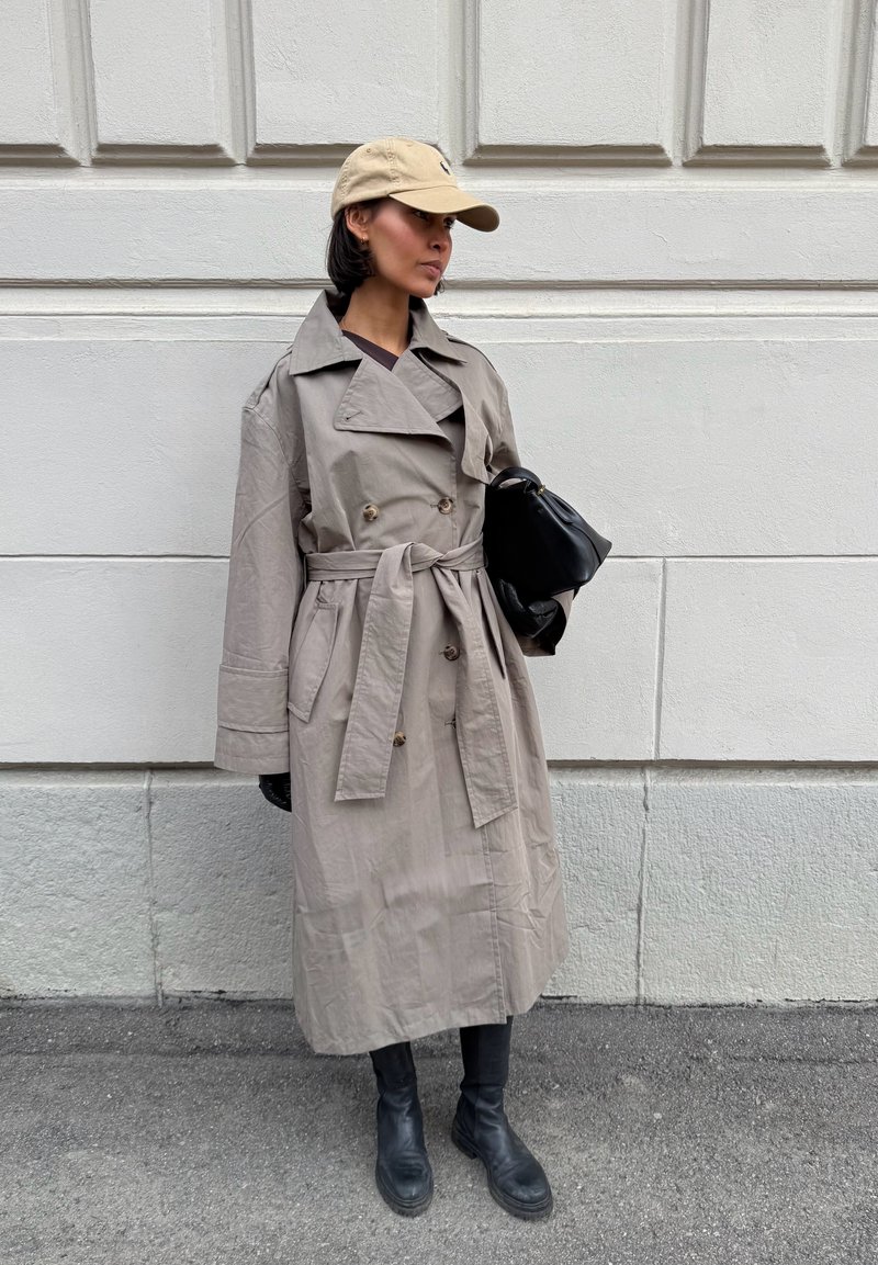 Femme portant un trench-coat beige, une casquette beige, des bottes noires, et tenant un sac à main noir, debout devant un mur gris clair.