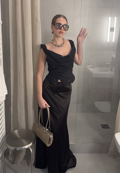 Mujer con top negro sin mangas y falda larga, gafas de sol grandes, collar de perlas, sosteniendo un bolso beige, de pie en un baño.