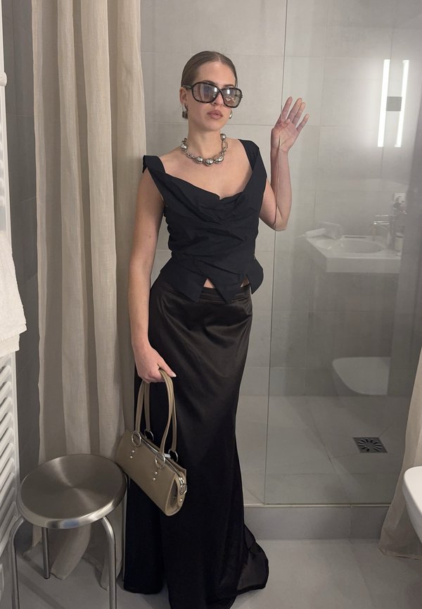 Frau trägt ein schwarzes ärmelloses Oberteil und einen langen Rock, übergroße Sonnenbrille, Perlenkette, hält eine beige Handtasche, steht in einem Badezimmer.