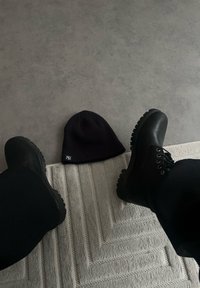 Bonnet en tricot noir avec un revers plié et un logo, posé sur un tapis clair texturé, à côté de bottines noires à lacets avec une semelle robuste.