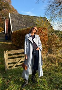 Jasnoniebieski płaszcz trencz z paskiem w talii, zestawiony z ciemnymi dżinsami i czarnymi kozakami do kolan; trzyma małą brązową torebkę na ramię.