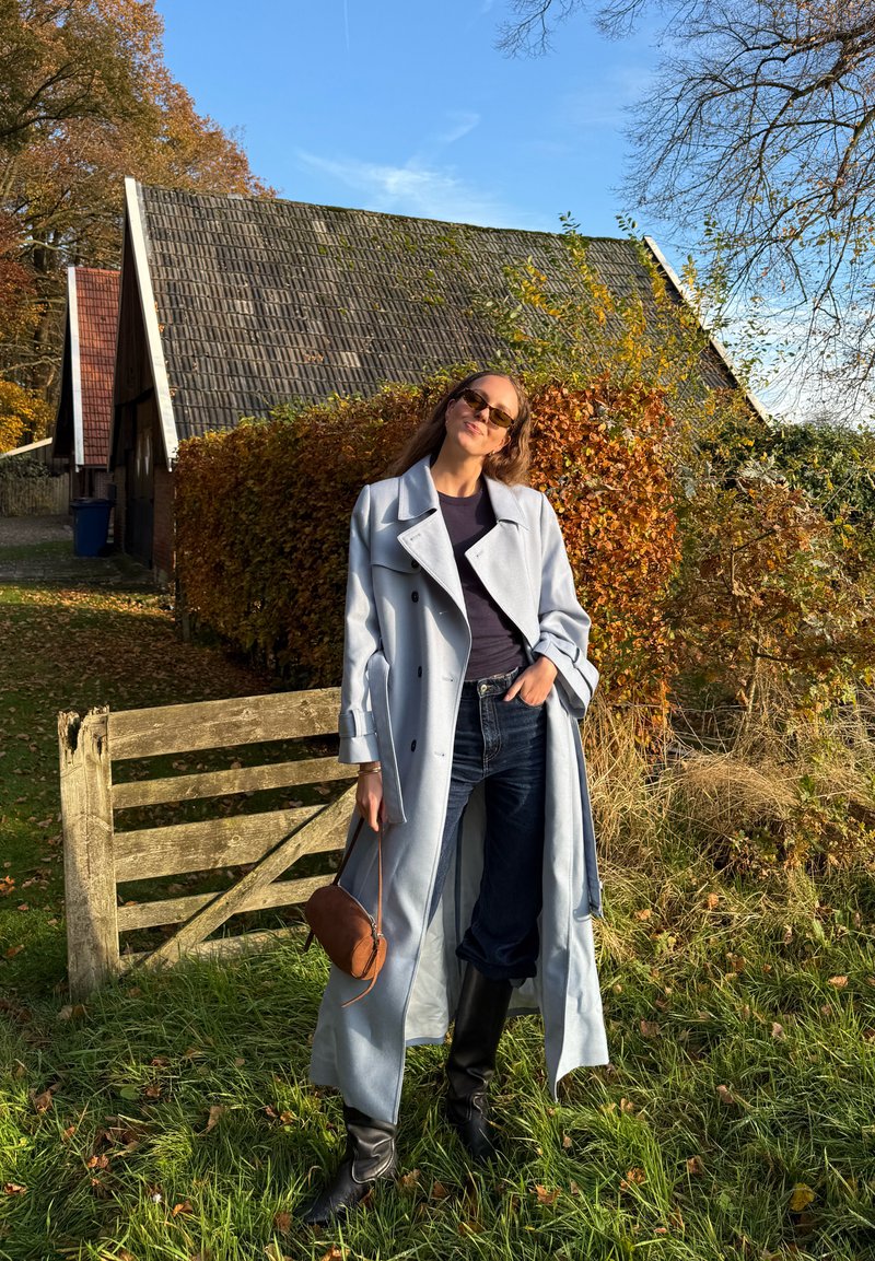 Trench azzurro chiaro con cintura in vita, abbinato a jeans scuri e stivali neri al ginocchio; porta una piccola borsa a tracolla marrone.