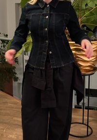 Schwarze Denimjacke mit Kragen, sichtbaren Nähten und silbernen Knöpfen. Kombiniert mit lockeren schwarzen Hosen mit gebundener Taillenakzentuierung.