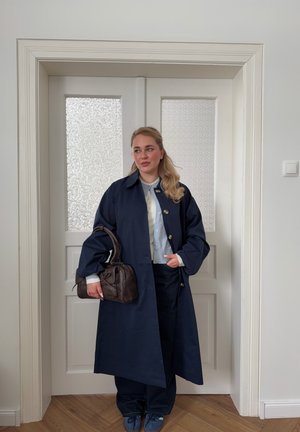 Mujer con un abrigo largo azul marino sosteniendo un bolso marrón oscuro, de pie frente a unas puertas dobles blancas con paneles de vidrio texturizado.