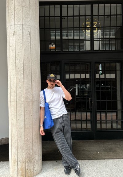 Una persona vestida con una camiseta blanca y pantalones a rayas se apoya en una columna, sosteniendo unas gafas de sol. Una bolsa de lona azul descansa sobre su hombro.