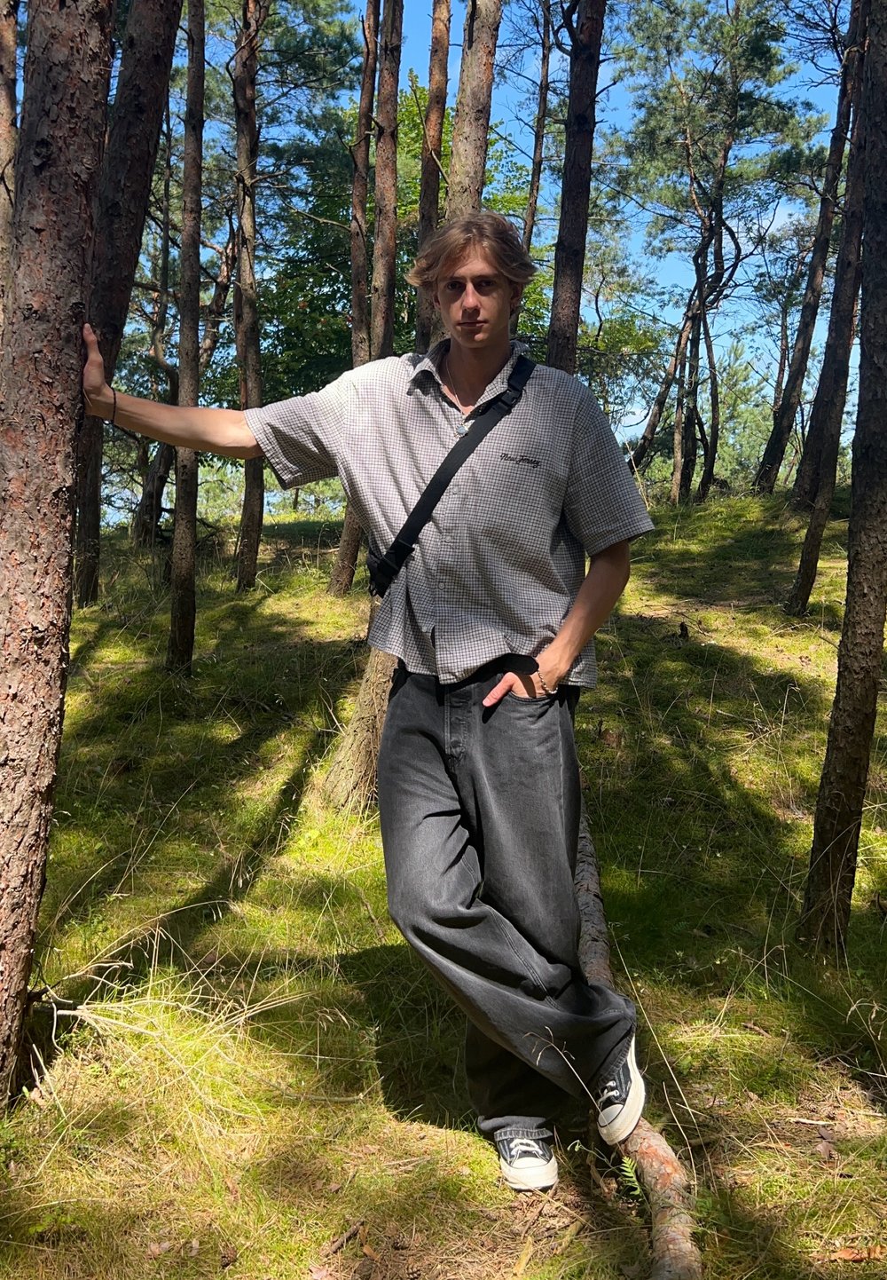 Persona con capelli corti che indossa una camicia a scacchi e jeans scuri, in piedi contro un albero in una zona boschiva con muschio verde.
