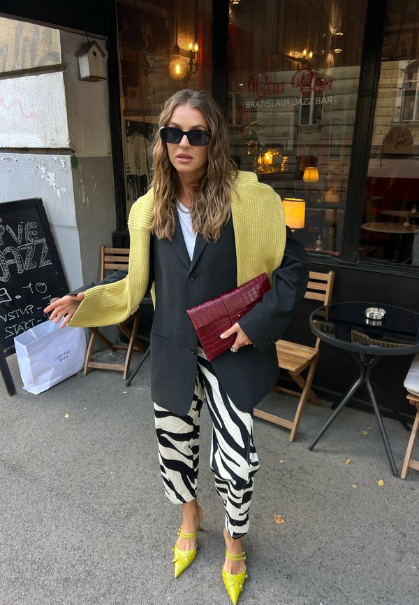 Mujer con blazer negro oversize, pantalones con estampado de cebra, suéter amarillo sobre los hombros, tacones amarillos, gafas de sol negras, sosteniendo un bolso rojo fuera de una cafetería.