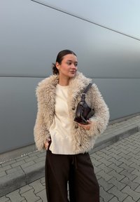 Faux fur jacket in light beige with a shaggy texture, paired with a cream jumper and brown wide-leg trousers. Holding a small dark brown bag.