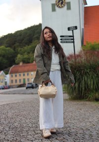 Veste vert olive portée sur un pull en maille blanc, pantalon blanc large, baskets beiges, et sac à main texturé beige avec une anse ronde.