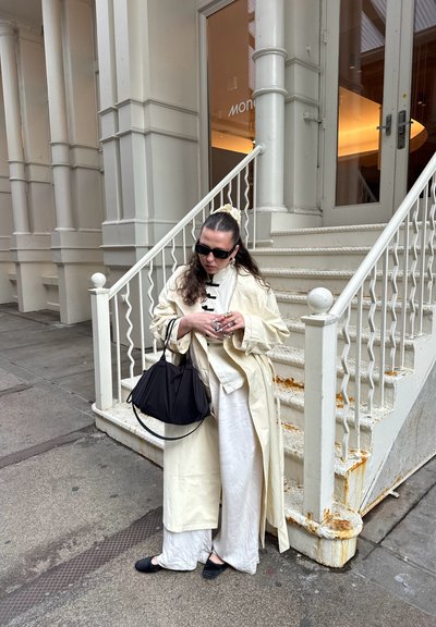 Mujer con gabardina ligera y gafas de sol sostiene un bolso negro, de pie cerca de una escalera blanca oxidada frente a un edificio beige.