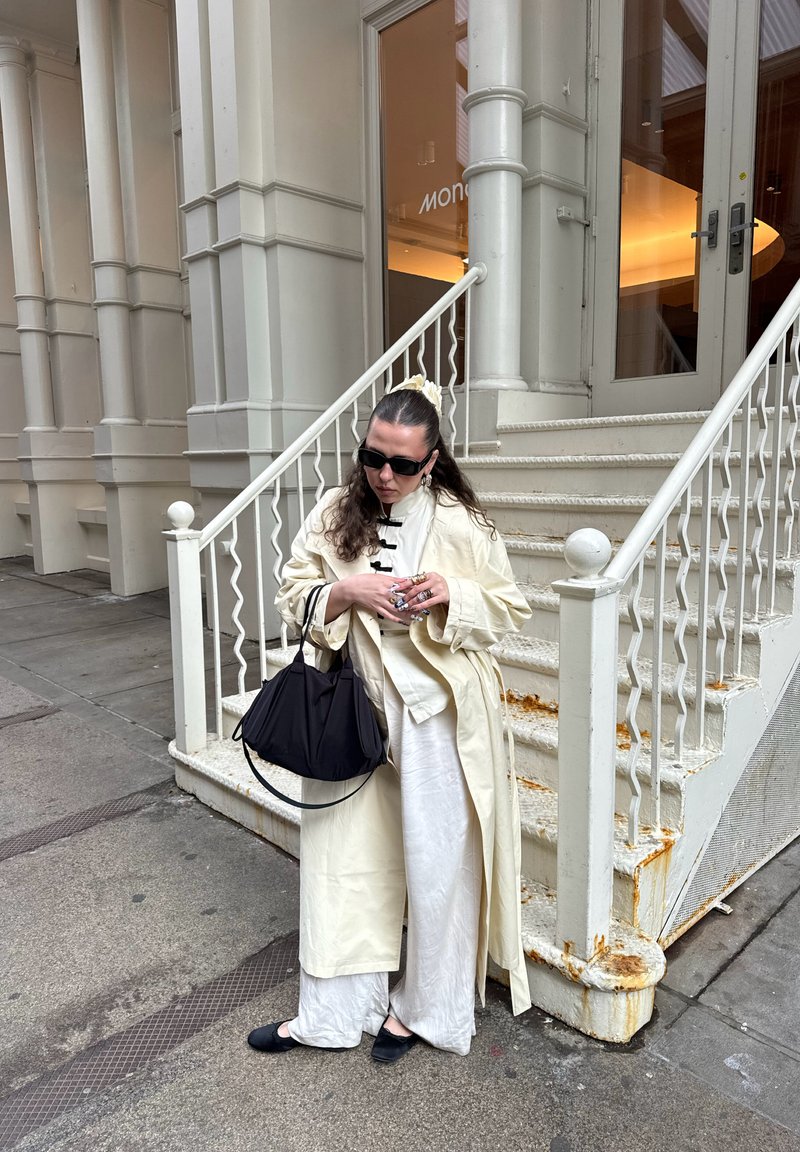 Femme portant un trench-coat léger et des lunettes de soleil, tenant un sac noir, debout près d'un escalier blanc rouillé à l'extérieur d'un bâtiment beige.