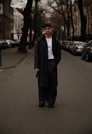 Abrigo largo negro, camiseta blanca y pantalones negros holgados. De pie en una calle con árboles, llevando zapatos negros y sosteniendo gafas de sol.