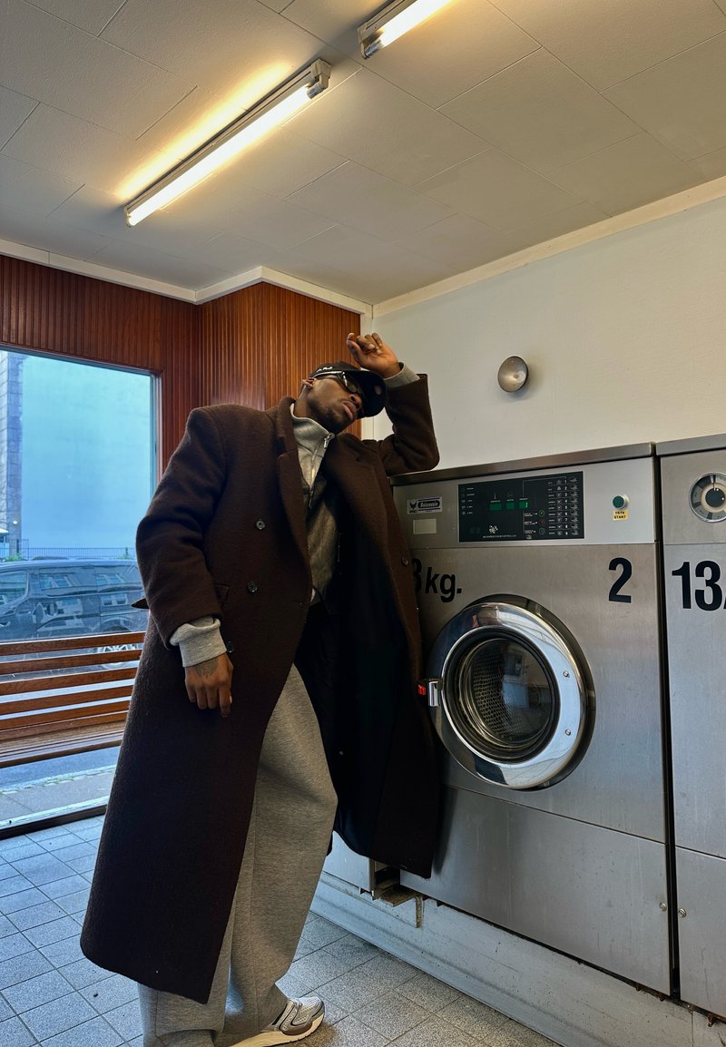 Homme en long manteau sombre et pantalon de survêtement gris appuyé contre une machine à laver à l'intérieur d'une laverie avec un banc en bois et une grande fenêtre.