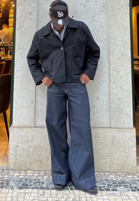 Crna kratka jakna s dva prednja džepa, uparena s tamnim trapericama širokih nogavica i crnom kapom. Glatka tekstura i suzdržan dizajn.