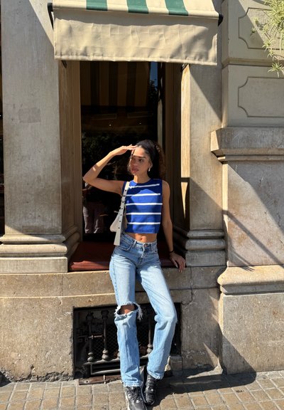 Top corto de rayas azul y blanco, jeans azules desgastados con rodilleras rasgadas, zapatillas negras y bolso blanco, posando frente a un muro de piedra.