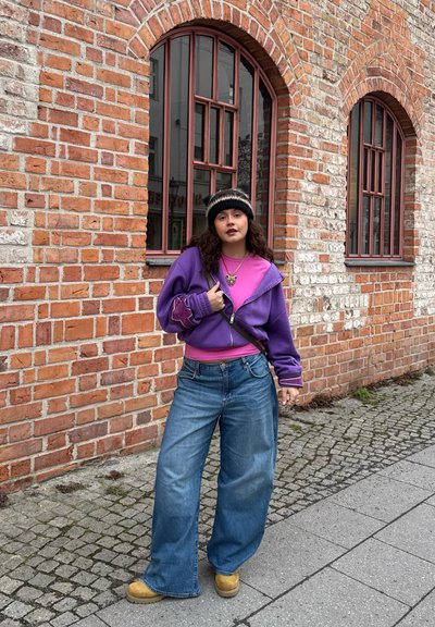 Joven mujer con chaqueta morada, camiseta rosa, pantalones vaqueros anchos y gorro de punto, de pie en una calle empedrada junto a un edificio de ladrillo con ventanas arqueadas.
