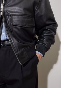 Κοντινό πλάνο ενός ατόμου που φοράει μαύρο δερμάτινο μπουφάν με εμπρόσθιο φερμουάρ, τσέπη με καπάκι, μπλε ριγωτό πουκάμισο, μαύρη ζώνη και μαύρο παντελόνι.