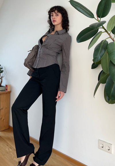 Camisa de manga larga a rayas en tonos neutros, pantalones negros ajustados y un bolso beis; está de pie contra una pared blanca con hojas verdes cerca.