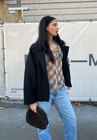 Casaco preto oversized, camisa de botão xadrez em bege e castanho, jeans de ganga azul claro e bolsa de mão castanha trançada, com cabelo longo e escuro.