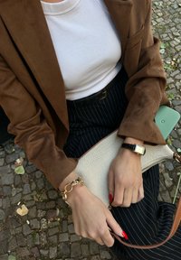 Bruin suède blazer over een witte geribbelde shirt, zwarte pinstripe broek, gouden armband en een beige clutch met een bruine afwerking.