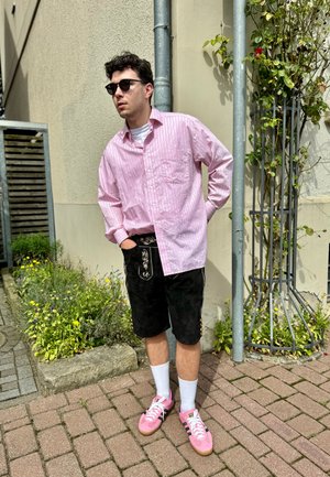 Camisa de rayas rosa claro de manga larga sobre una camiseta blanca; pantalones cortos negros con bordado; zapatillas rosas; gafas de sol negras, de pie contra una pared.