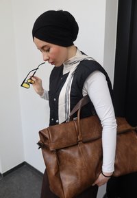 Femme portant un foulard noir et un gilet, tenant des lunettes de soleil et un grand sac en cuir marron à l'intérieur près d'un mur blanc.