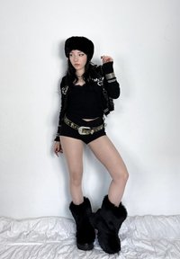Chapeau en fausse fourrure noire, haut moulant noir, short noir et ceinture ornée. Bottes noires en fourrure avec semelles épaisses, posées sur un drap blanc.