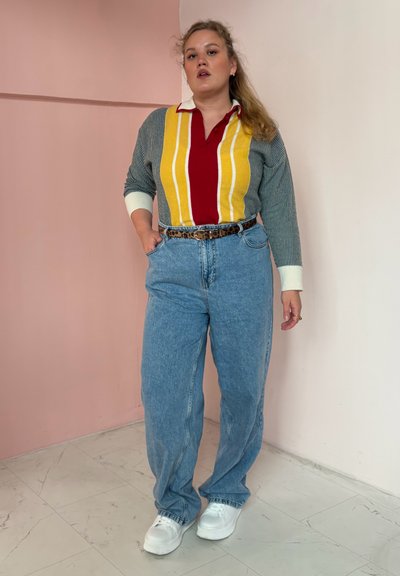 Persona de pie contra una pared bicolor, vistiendo una camiseta de rayas multicolor de manga larga, jeans azules con un cinturón de leopardo y zapatillas blancas.