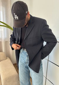 Blazer preto riscado usado sobre uma camisa preta, combinado com jeans azul claro. Um boné preto está usado e um anel de prata é visível.