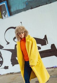 Casaco de fleece amarelo com grandes lapelas, usado sobre um suéter laranja. Um chapéu de pelúcia cinza e óculos estampados completam o visual contra um fundo de graffiti.