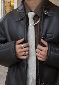 Fekete bőrkabát gombos részletekkel, barna ing és világos bézs nyakkendő fölött viselve. Több ezüst gyűrű és egy lánc emeli ki a megjelenést.