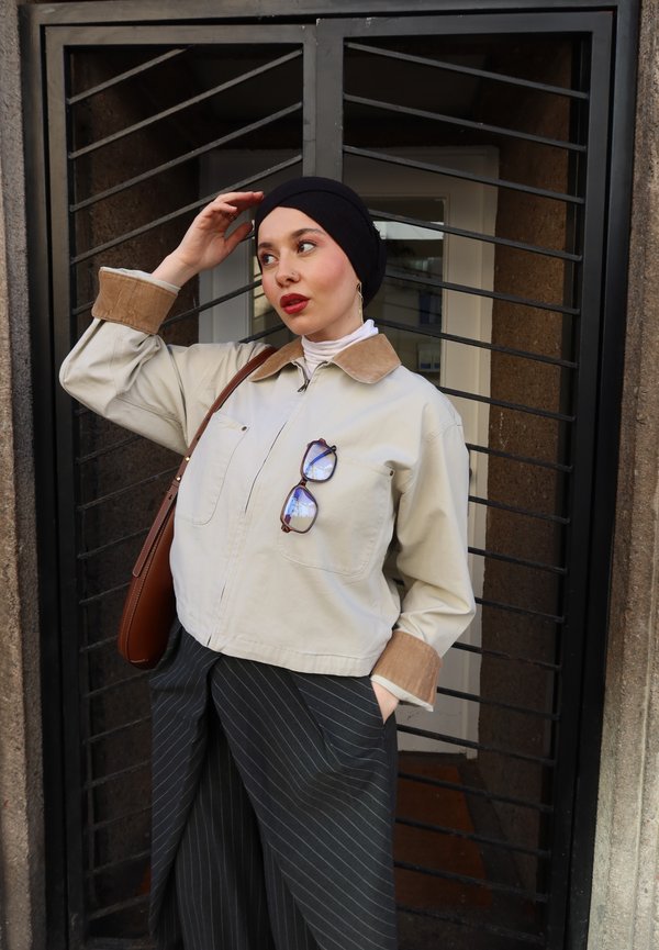 Frau mit schwarzem Kopftuch, beiger Jacke mit hängender Sonnenbrille, gestreifter Hose und brauner Umhängetasche steht vor einem Metalltor.
