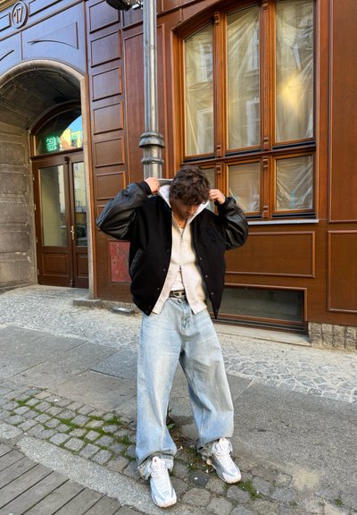 Chaqueta varsity negra con mangas de cuero, sudadera gris por debajo, jeans holgados de color azul claro y zapatillas blancas. Fondo de calle de adoquines.