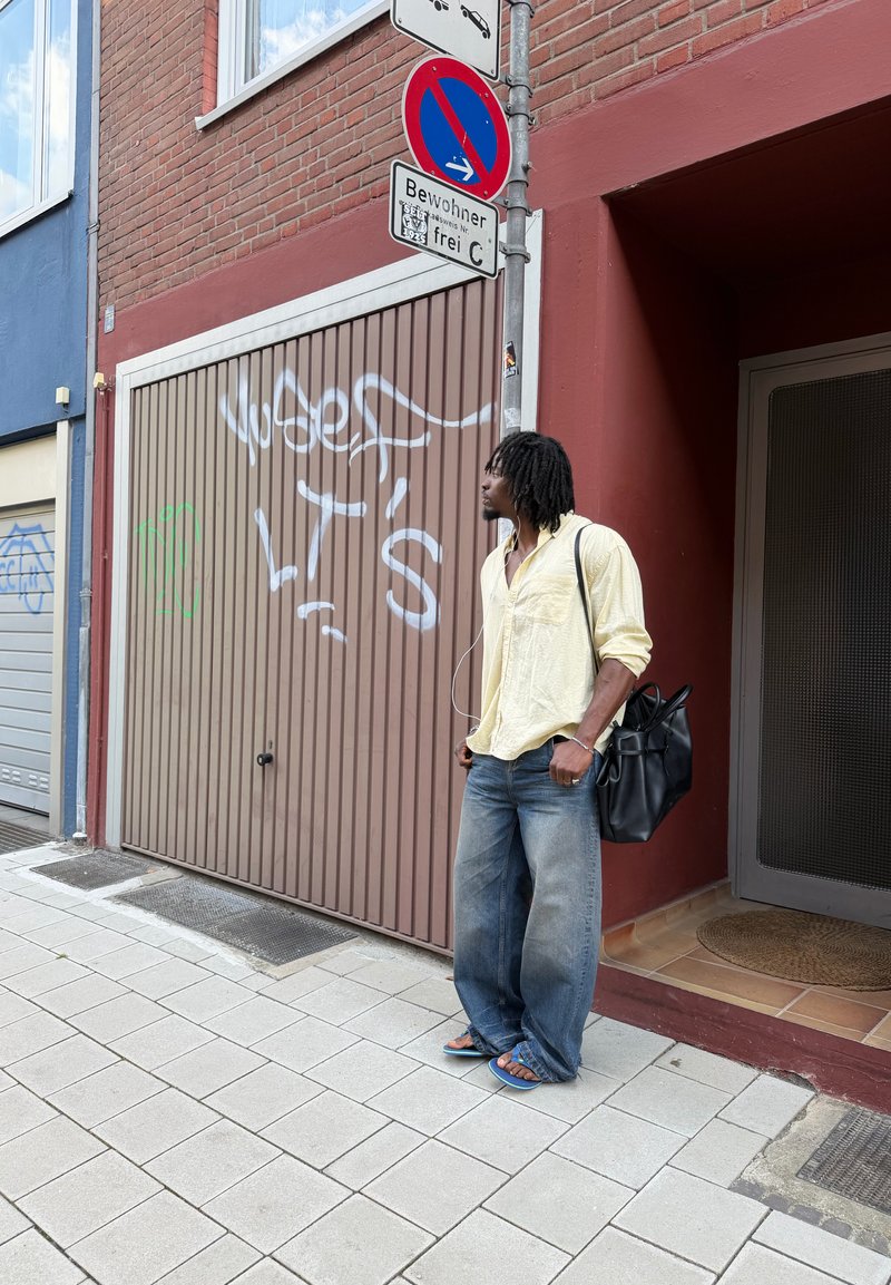 Uomo con camicia gialla, jeans e sandali in piedi sul marciapiede vicino a una porta del garage con graffiti e cartello di divieto di sosta sopra.