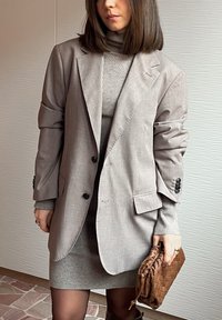 Femme portant un blazer gris clair oversize sur une robe col roulé côtelée, tenant une pochette marron texturée, debout contre un mur clair texturé.