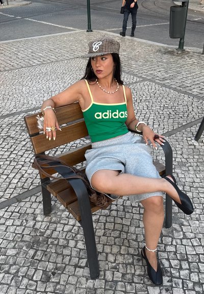 Top de tirantes verde y amarillo de Adidas, pantalones cortos grises, zapatos negros sin cordones, sentado en un banco de madera con un pavimento de piedra texturizada.