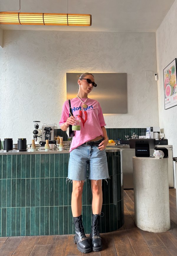 Femme portant un t-shirt graphique rose et un short en jean se tient dans un café moderne, tenant une boisson verte avec des machines à café derrière elle.