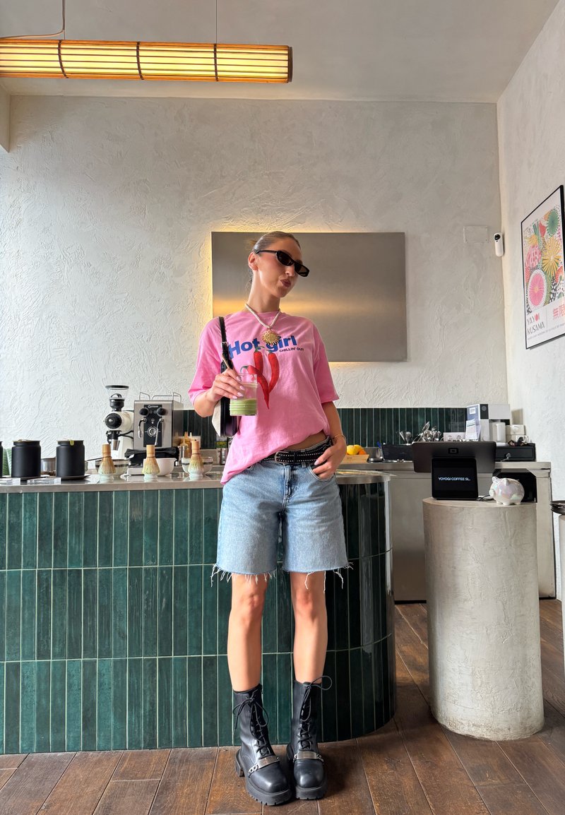 Femme portant un t-shirt graphique rose et un short en jean se tient dans un café moderne, tenant une boisson verte avec des machines à café derrière elle.