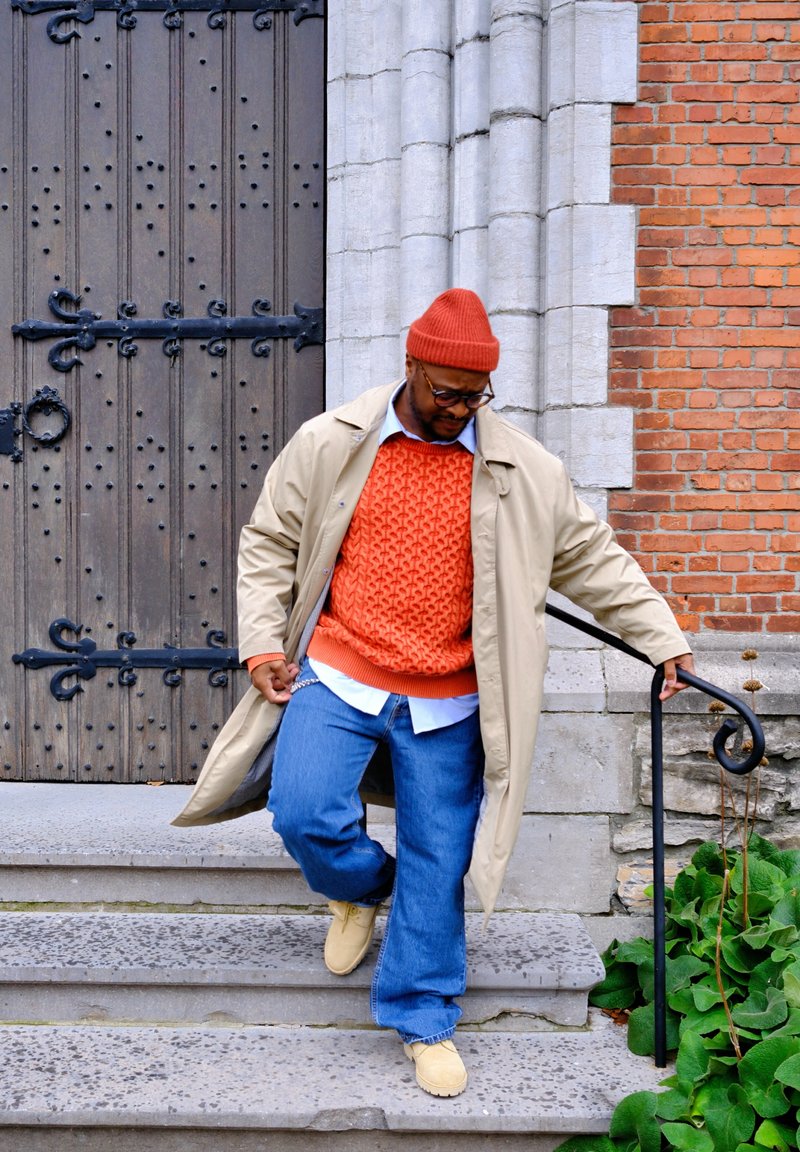 Cappotto impermeabile beige, maglione arancione a trama, jeans blu, scarpe beige. Persona che scende scale di pietra vicino a una grande porta di legno con accenti in metallo.